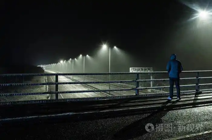 一个孤独的戴着兜帽的身影站在桥上晚上在暴风雪中俯视一条空荡荡的
