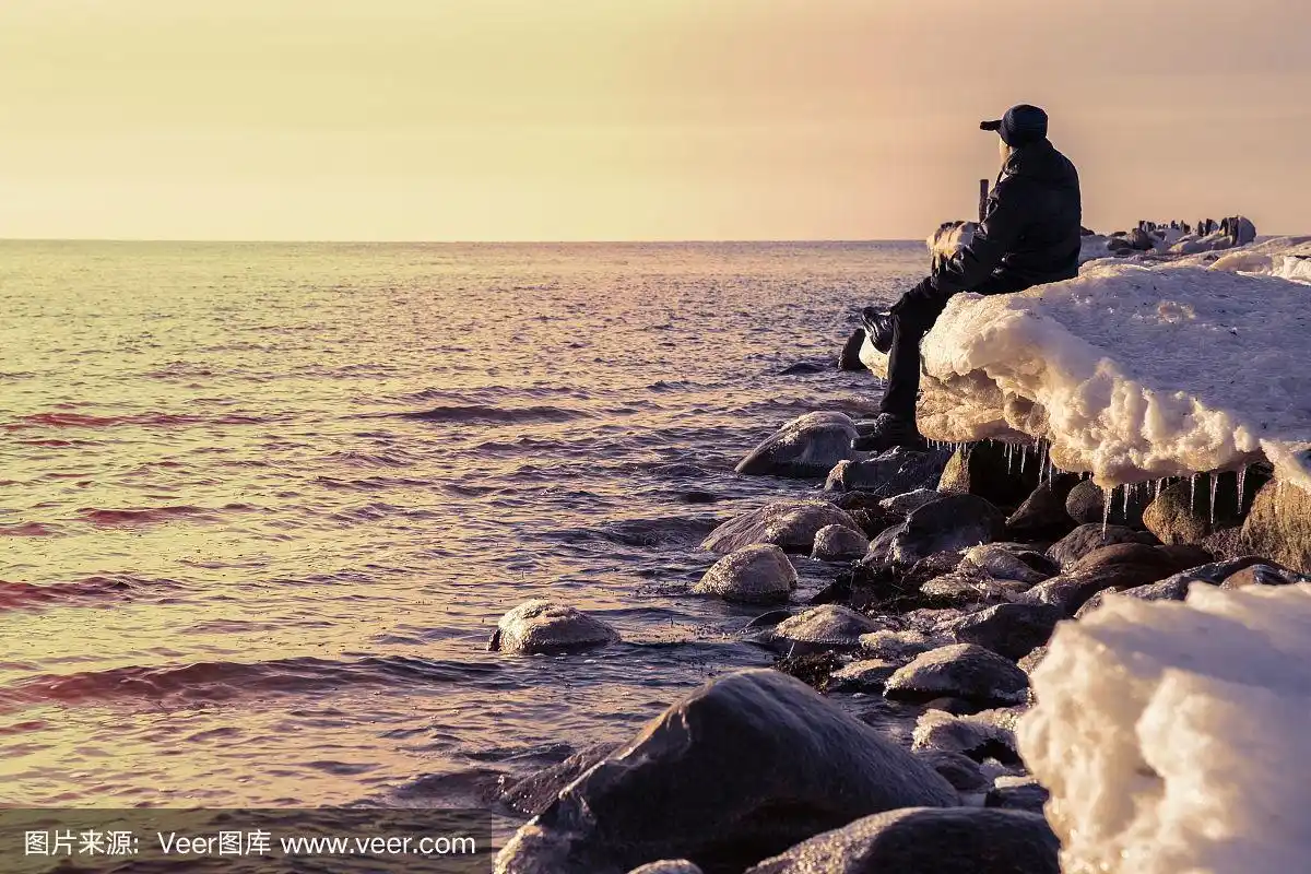 年轻的成年人独自坐在海边旧防波堤上覆盖着雪的岩石上.