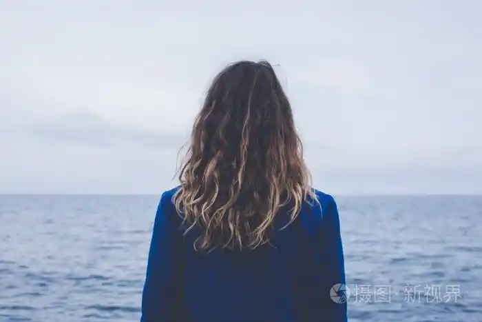 金发卷曲女孩在看着朦胧的阳光透过厚厚的雾平静的大海和蓝天的背影