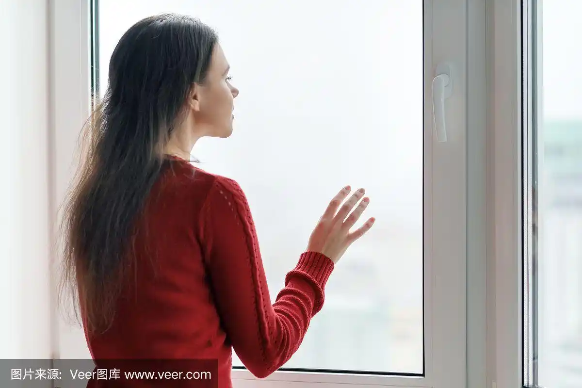 穿着红色毛衣的年轻女子望着窗外,女性把她的手放在窗户玻璃上,侧视图