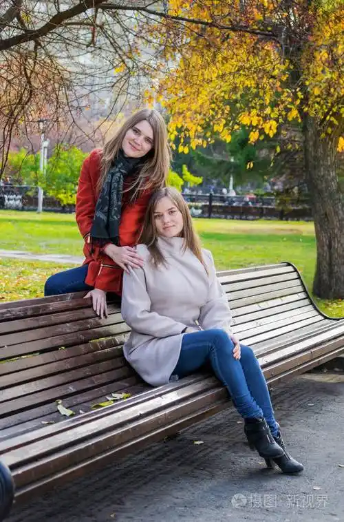 两个美丽的女朋友在阳光明媚的秋日并排坐在长凳上微笑