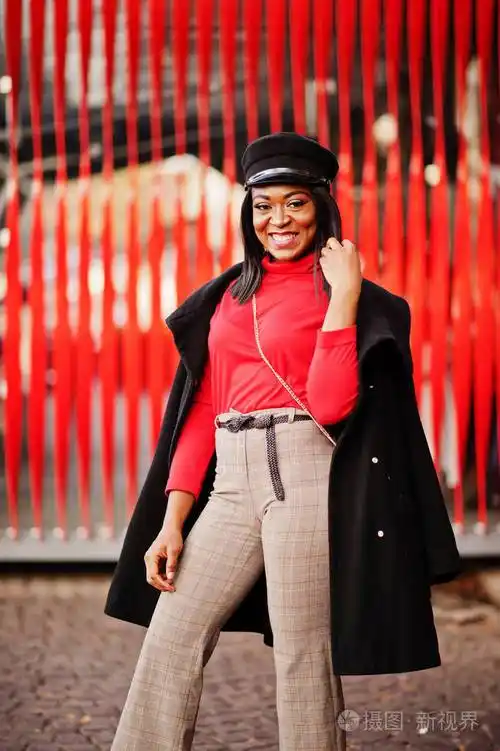 穿着外套和报童帽的非裔美国人时尚女孩在红色背景下在街上摆姿势