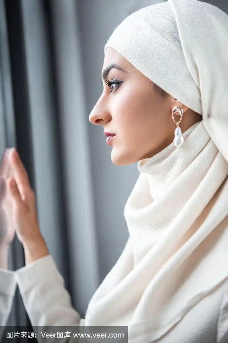 年轻漂亮的穆斯林妇女望着窗户的侧视图