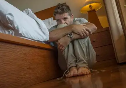 年轻的沮丧绝望的男人坐在卧室的地板上,挨着床,饱受抑郁和焦虑的折磨