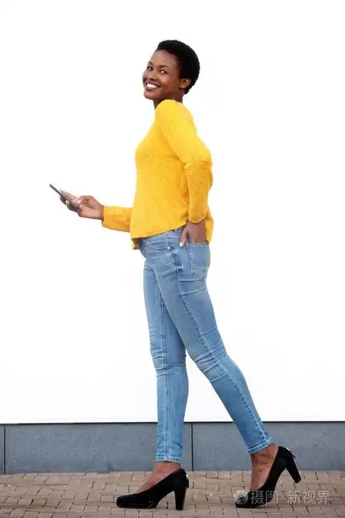 年轻的非裔美国妇女用白色墙壁用手机走路的全身侧面肖像