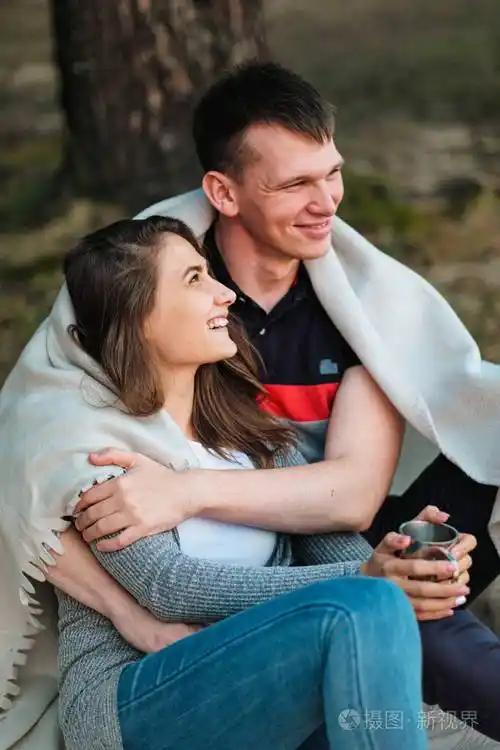 夕阳下一对相爱的情侣坐在河岸上他们拥抱在一起卷成格子花纹喝着金属