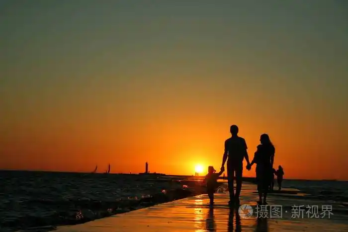 一家三口在堤坝上行走的剪影双手握着明亮的夕阳背景是晴朗的天空