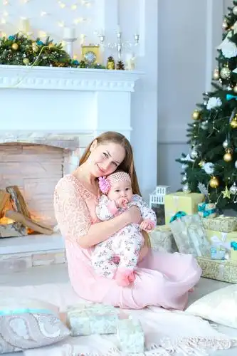 年轻的母亲抱着女儿在胳膊附近装饰的壁炉和圣诞树穿着粉红色的礼服