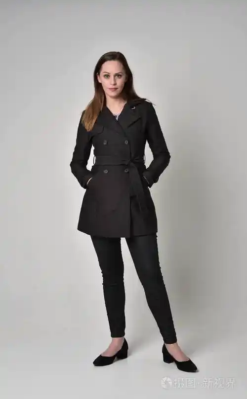 一个黑发女孩穿着黑色长外套站在灰色工作室背景上的姿势的全长肖像