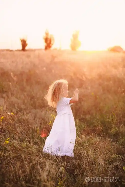 可爱的快乐的小女孩卷曲的金发穿着白色的礼服站在阳光灿烂的夕阳田野