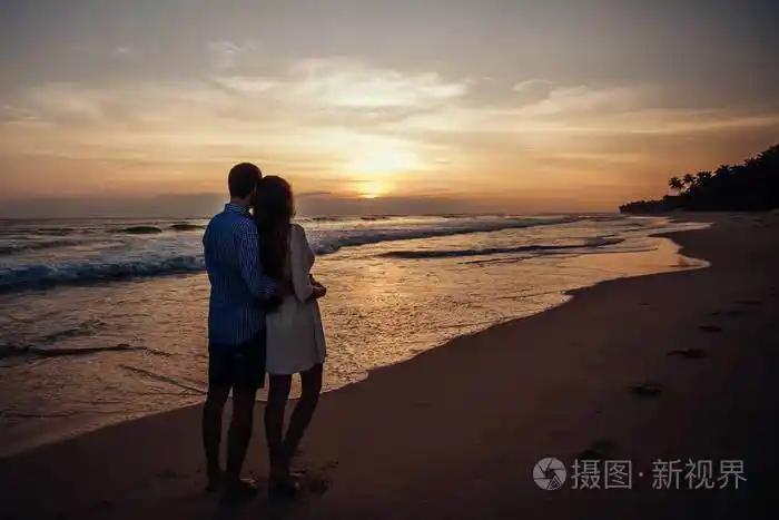 女孩和男孩的后视镜互相拥抱年轻夫妇在海滩上欣赏日落的背景五颜六色