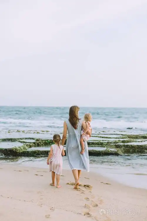 年轻的母亲与可爱的小女儿漫步在沙滩上后视图