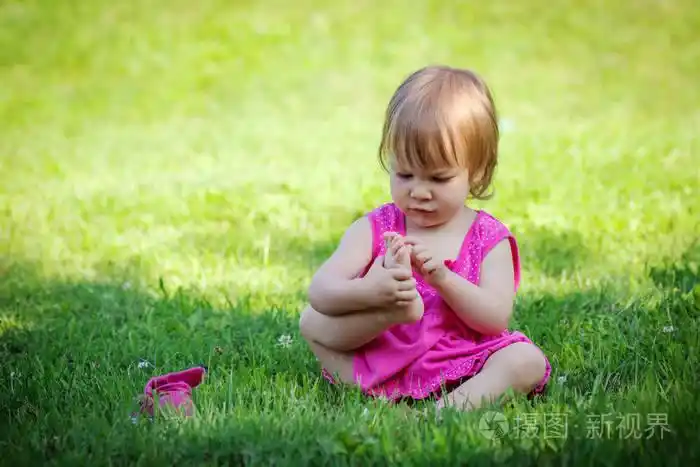 穿着粉红色连衣裙的可爱小女孩正在草地上玩她的脚趾 otdoors.