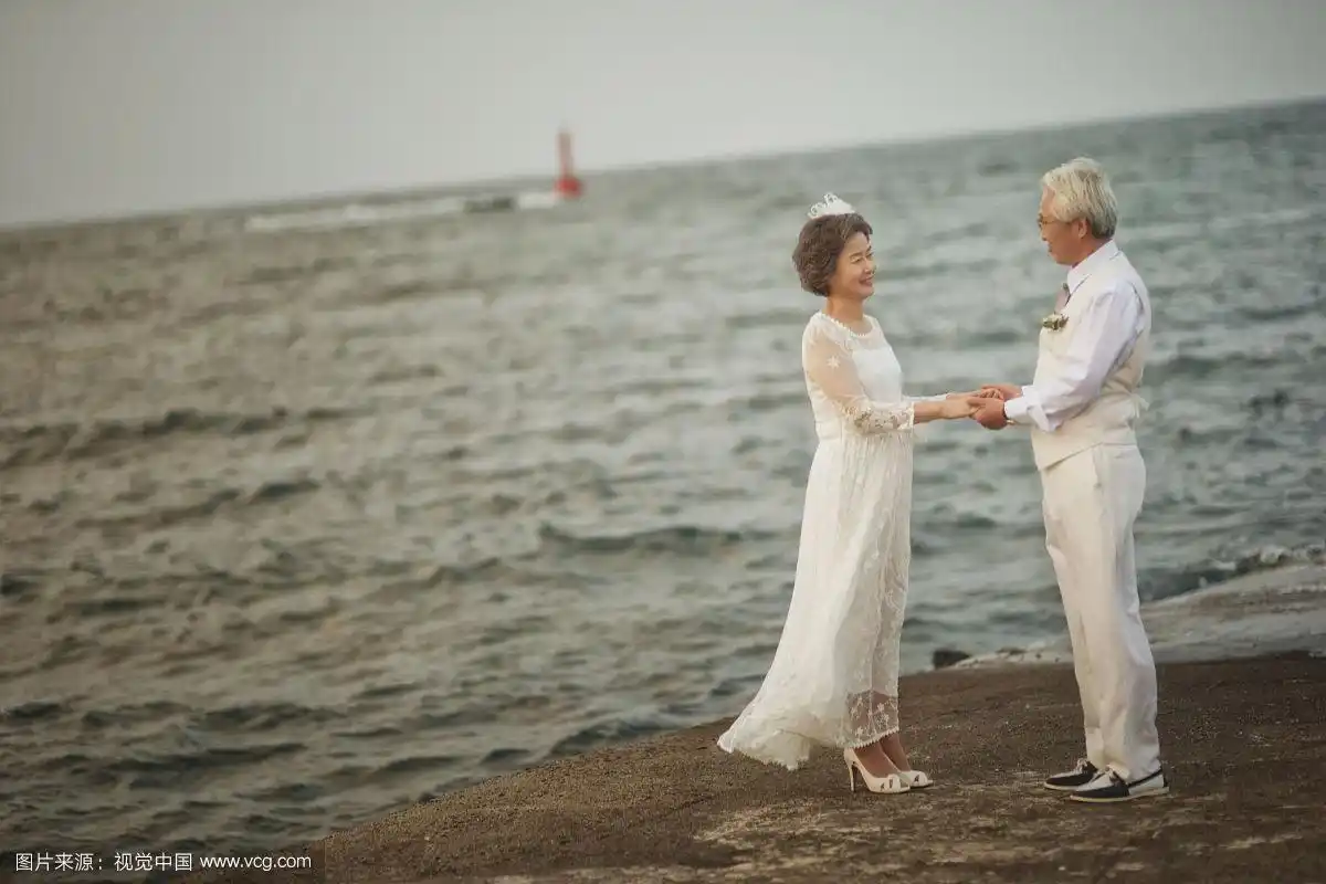 老夫妇穿着婚礼的样子手牵手面对面站在海滩上的照片