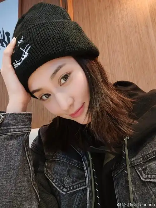 是棕黄色的,穿黑色卫衣搭配同色牛仔外套,头戴毛线帽,歪头微笑看向镜