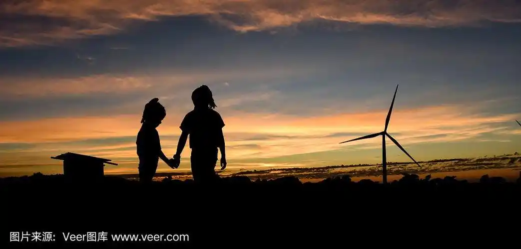 在夕阳的背景下,两个小女孩手牵手在风力涡轮机场的剪影.