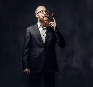 红发的大胡子男性身着西装和眼镜吸烟传统管道在深灰色背景照片