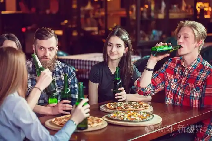 朋友们在酒吧里喝酒,他们坐在一张木桌旁,桌上摆着啤酒和披萨.