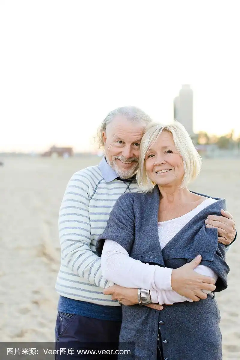 老丈夫拥抱微笑的妻子在沙滩的背景