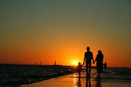 一家三口在堤坝上行走的剪影,双手握着明亮的夕阳,背景是晴朗的天空.