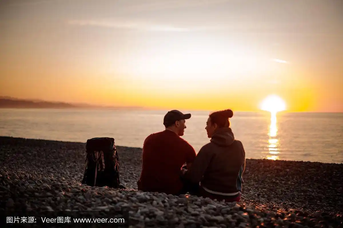 两个背着背包的徒步旅行者坐在海滩上看海