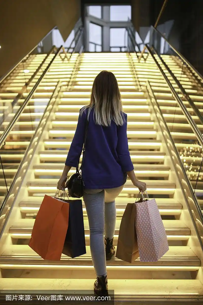 在购物中心的灯光楼梯上,年轻女子拿着购物袋.阅读观点