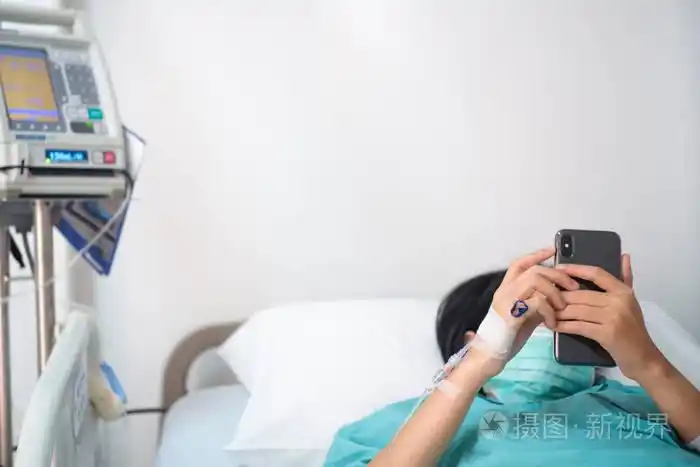 年轻妇女患者躺在床上在医院玩智能手机与静脉正常 salin照片-正版
