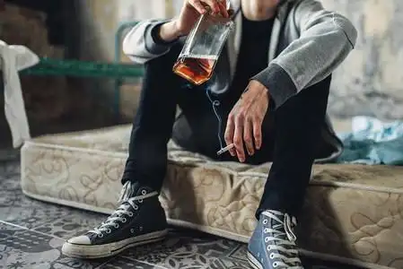 上瘾的男性坐在床垫上,手里拿着香烟和一瓶酒,背景是垃圾房内部.