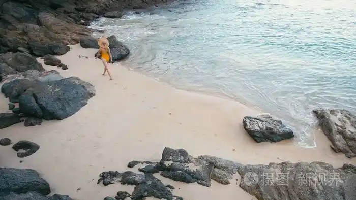 穿着黄色泳衣走在美丽的热带白色沙滩上的年轻女子的背影照片-正版